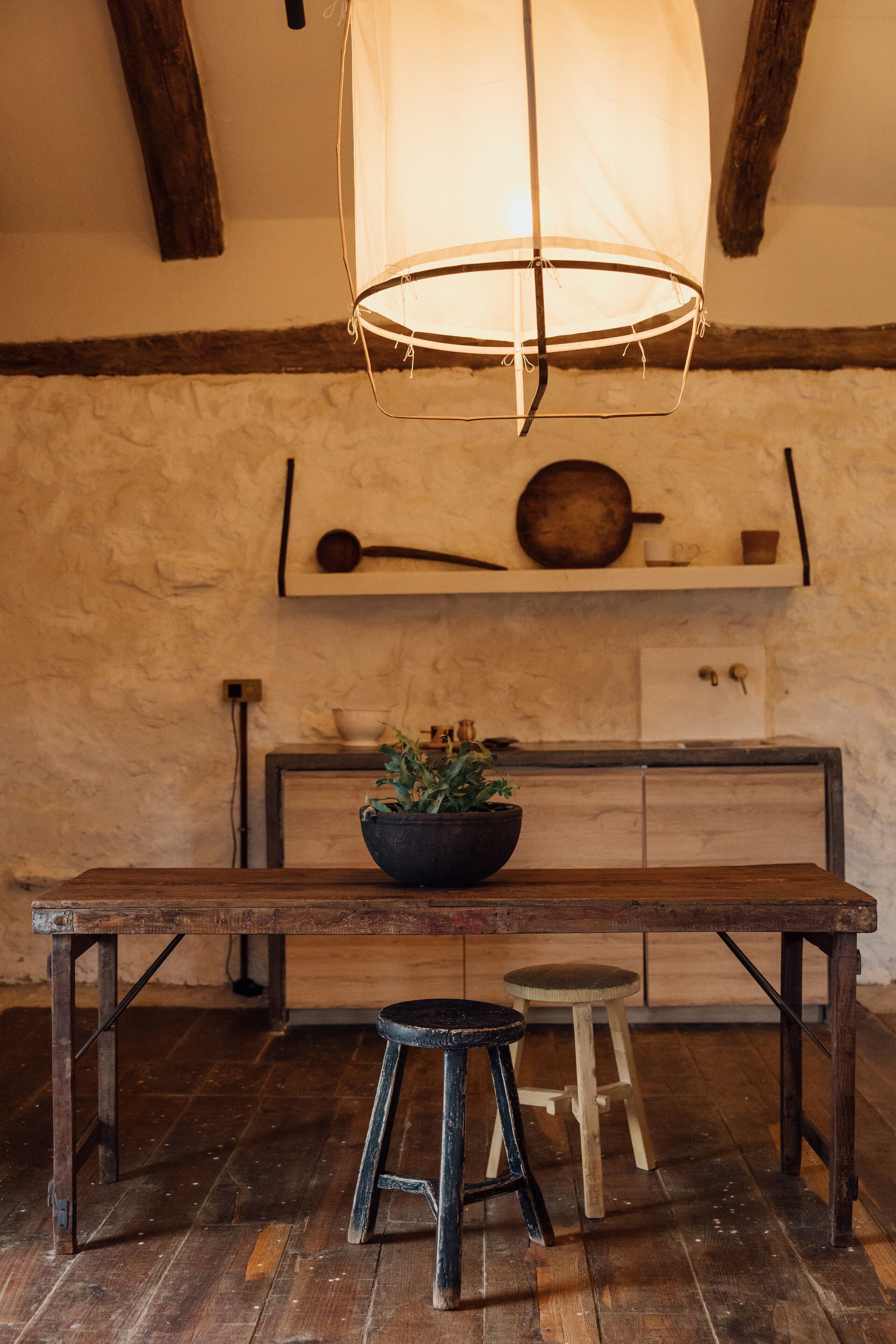 Een rustieke houten tafel met een plantenkom, twee krukken en een hanglamp in een ruimte met zichtbare balken en een plank met keukenbenodigdheden.