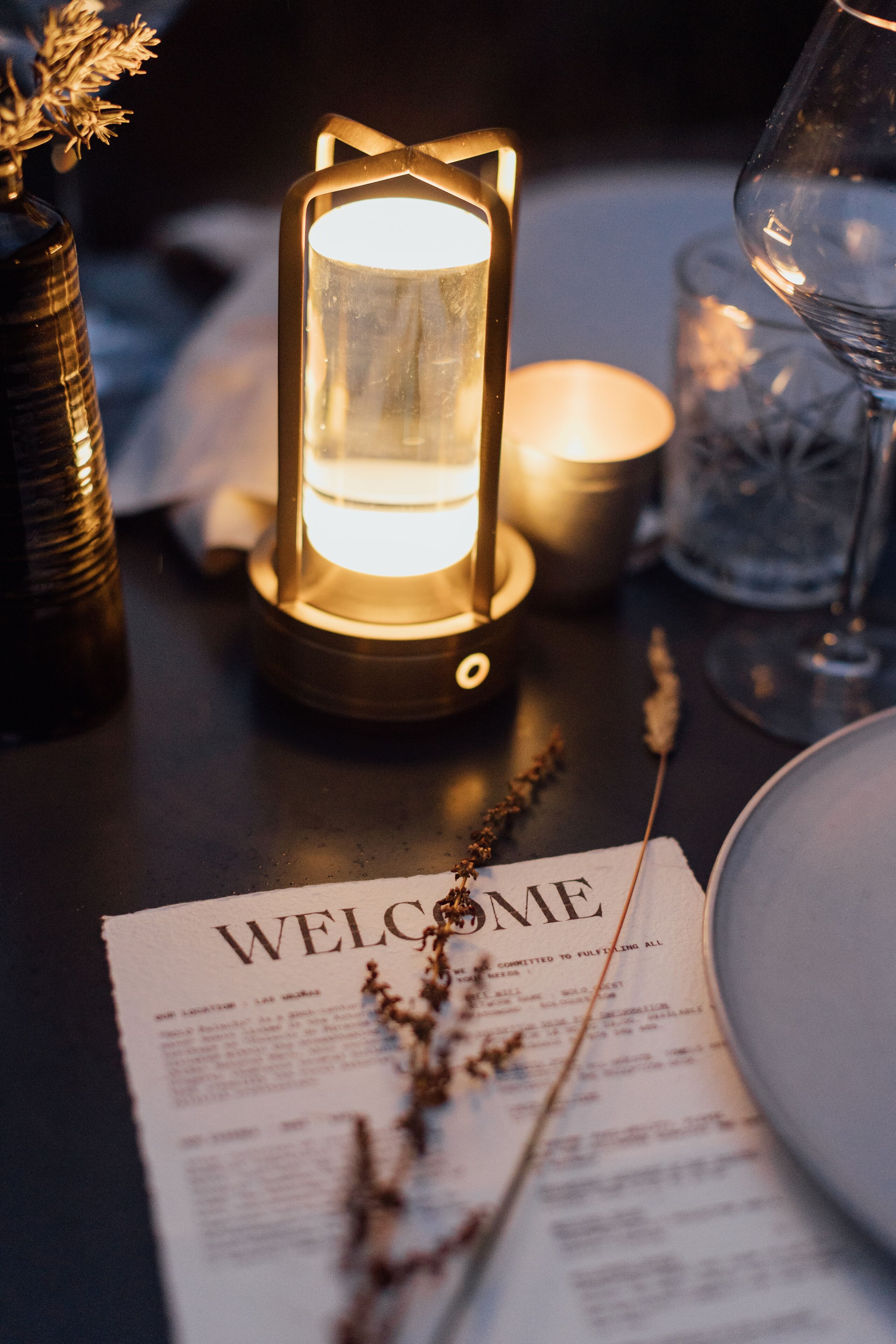 A table with a lit lantern, a glass of water, a candle, and a welcome sign.