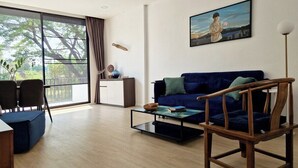 A modern living room with a blue sofa, a wooden chair, a glass coffee table, and a painting on the wall. - Your Local Spacious 3-story Home, Rooftop views & King Beds near Night Bazaar (Chang Wat Chiang Mai)