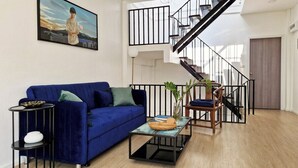 A modern living room with a blue sofa, a glass coffee table, and a staircase with a black railing. - Your Local Spacious 3-story Home, Rooftop views & King Beds near Night Bazaar (Chang Wat Chiang Mai)