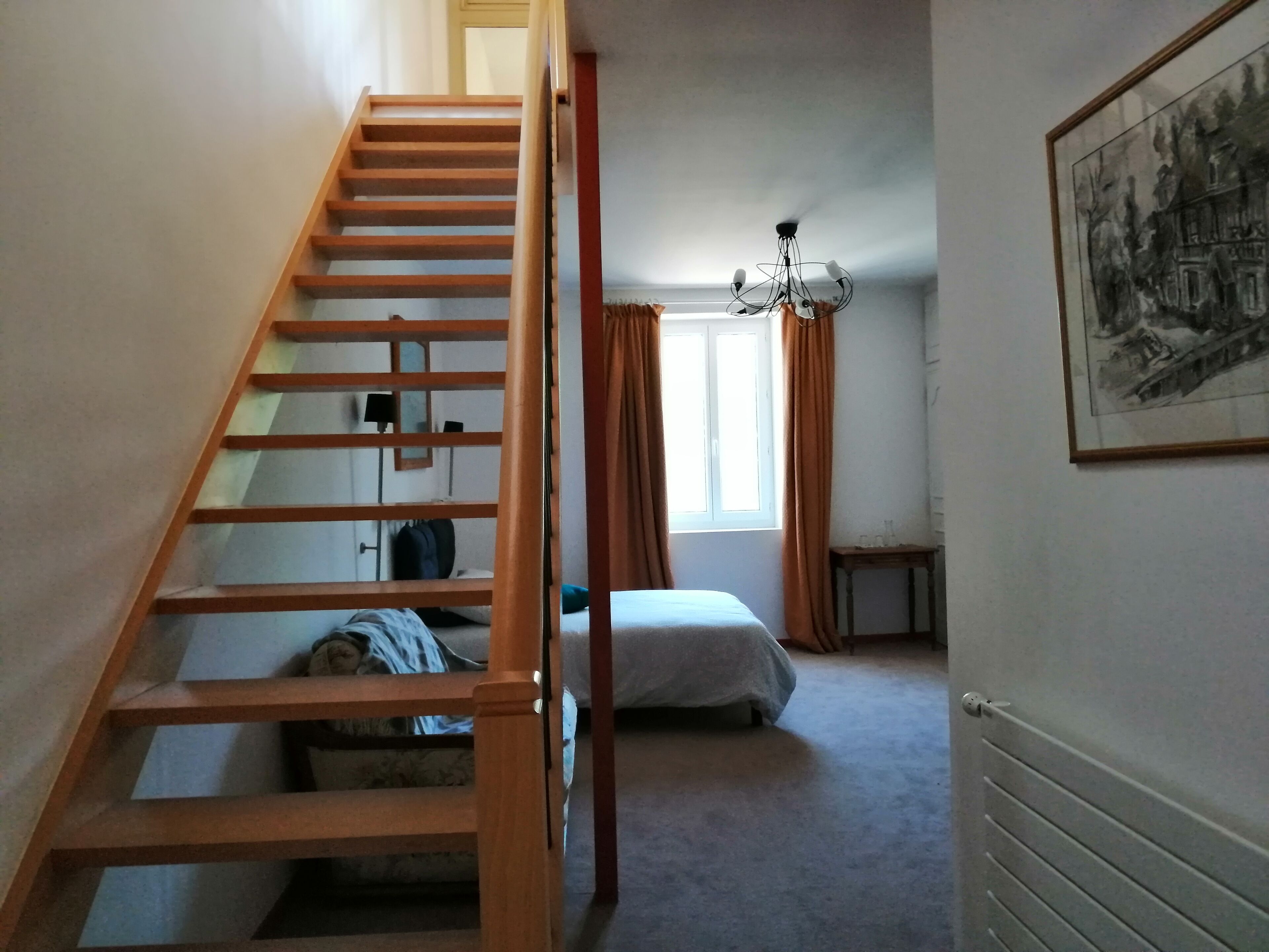 A bedroom with a wooden staircase, a bed, a window with curtains, a chandelier, and a radiator.