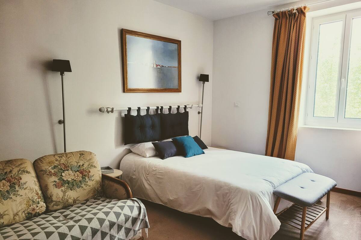 A bedroom with a bed, a nightstand, a chair, a window with curtains, and a wall-mounted lamp.