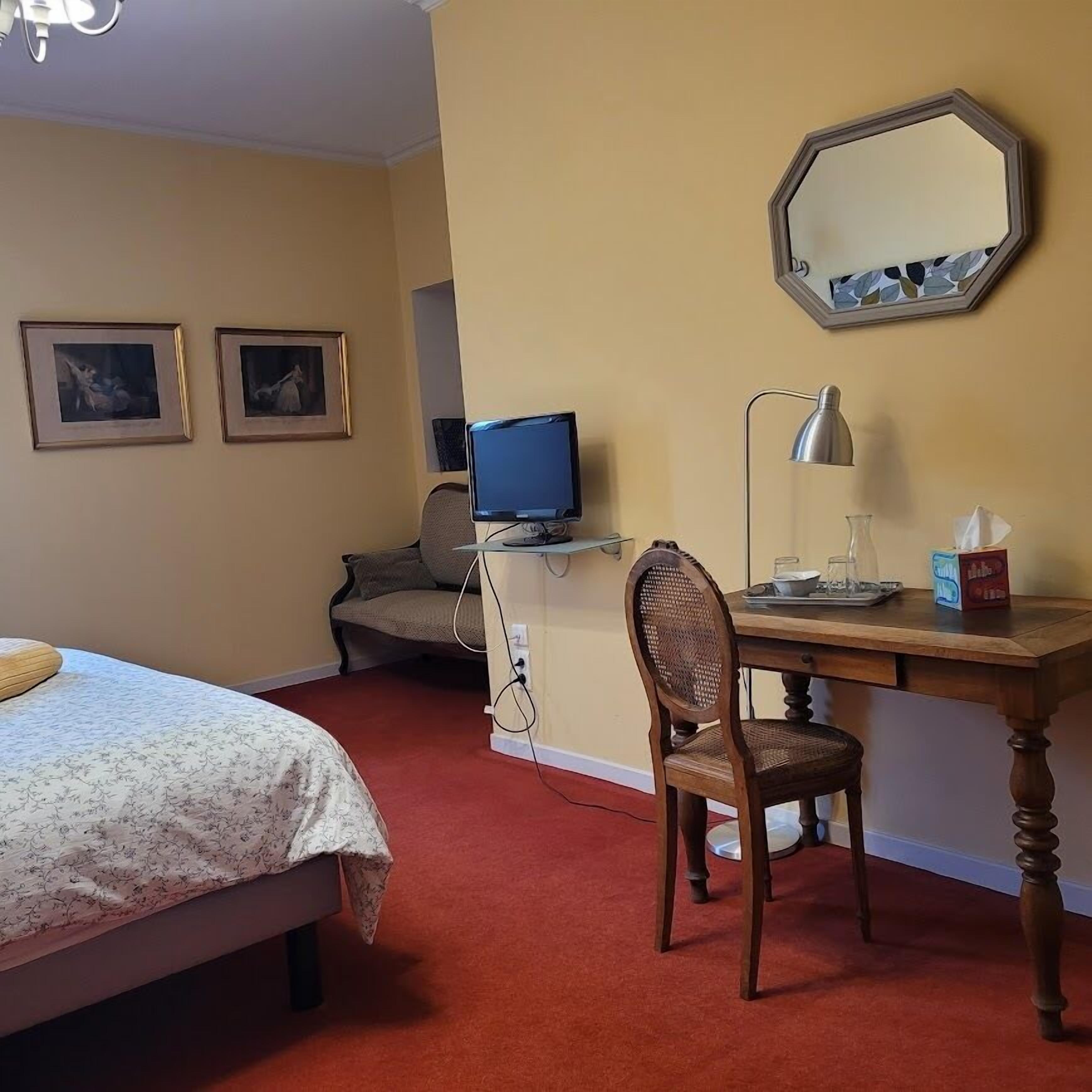 Ein Hotelzimmer mit einem Bett, einem Schreibtisch mit Computer, einem Stuhl, einer Schreibtischlampe und zwei gerahmten Bildern an der Wand.