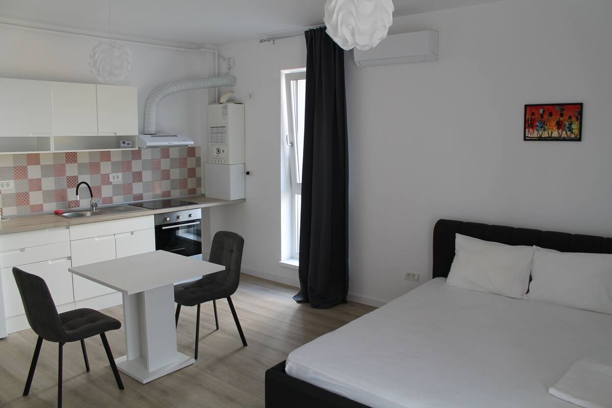 A modern hotel room with a bed, a small dining table with chairs, a kitchen area with a stove, and a window with curtains.
