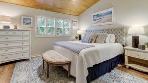 Ein Schlafzimmer mit Holzbalkendecke, einem großen Bett mit einem gepolsterten Kopfteil, einer Bank, einer Kommode und einem Fenster mit Jalousien.