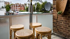 Un balcon doté d’une table en verre et de quatre chaises en osier, offrant une vue sur un quartier résidentiel avec ses maisons et une rivière.