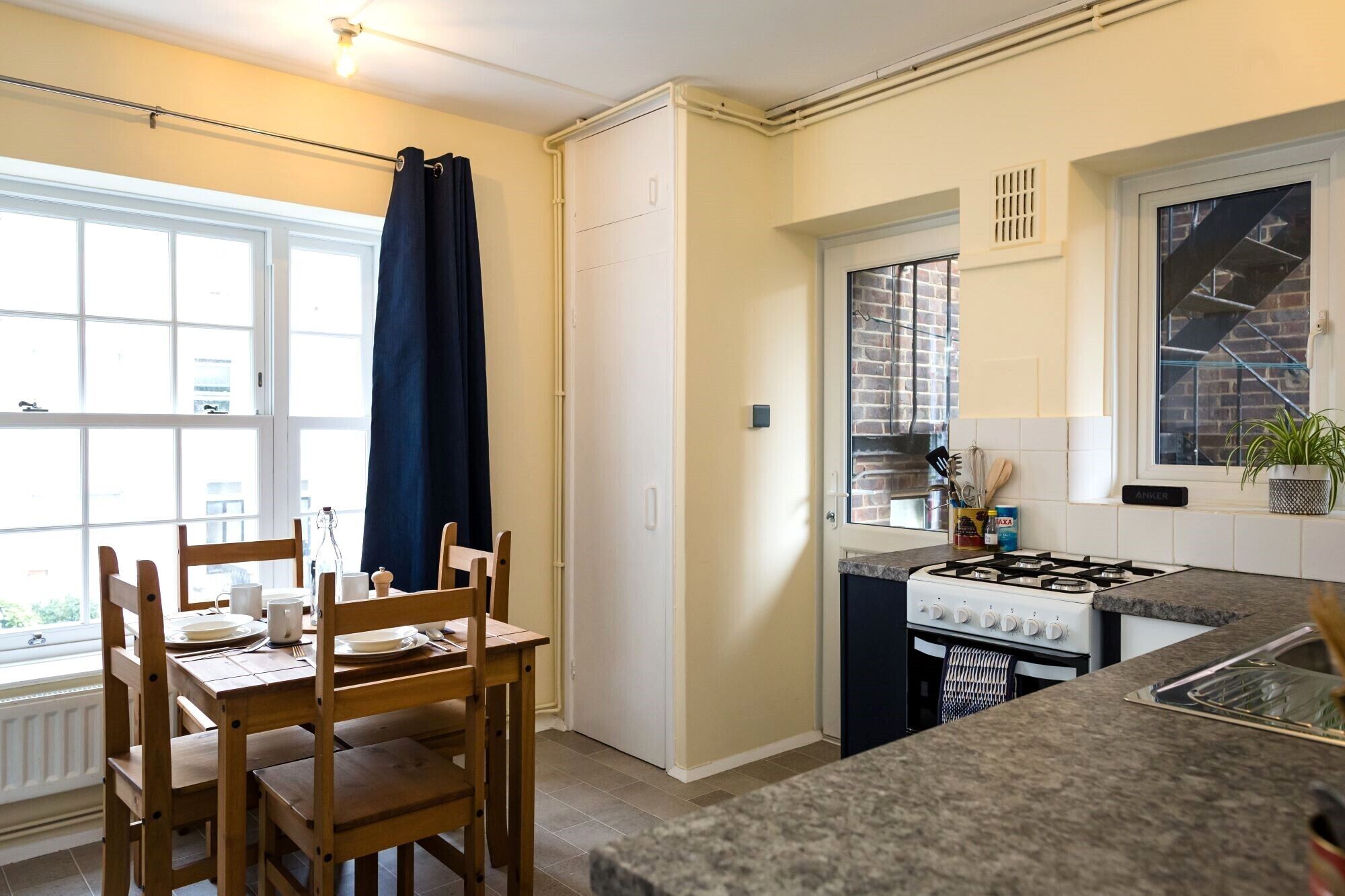 A compact kitchen with a gas stove, a window with a view of a brick building, and a dining area with wooden chairs and a table.