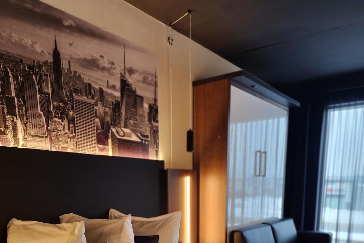 A hotel room with a large bed, a cityscape mural on the wall, a modern sofa, and a window with blinds.