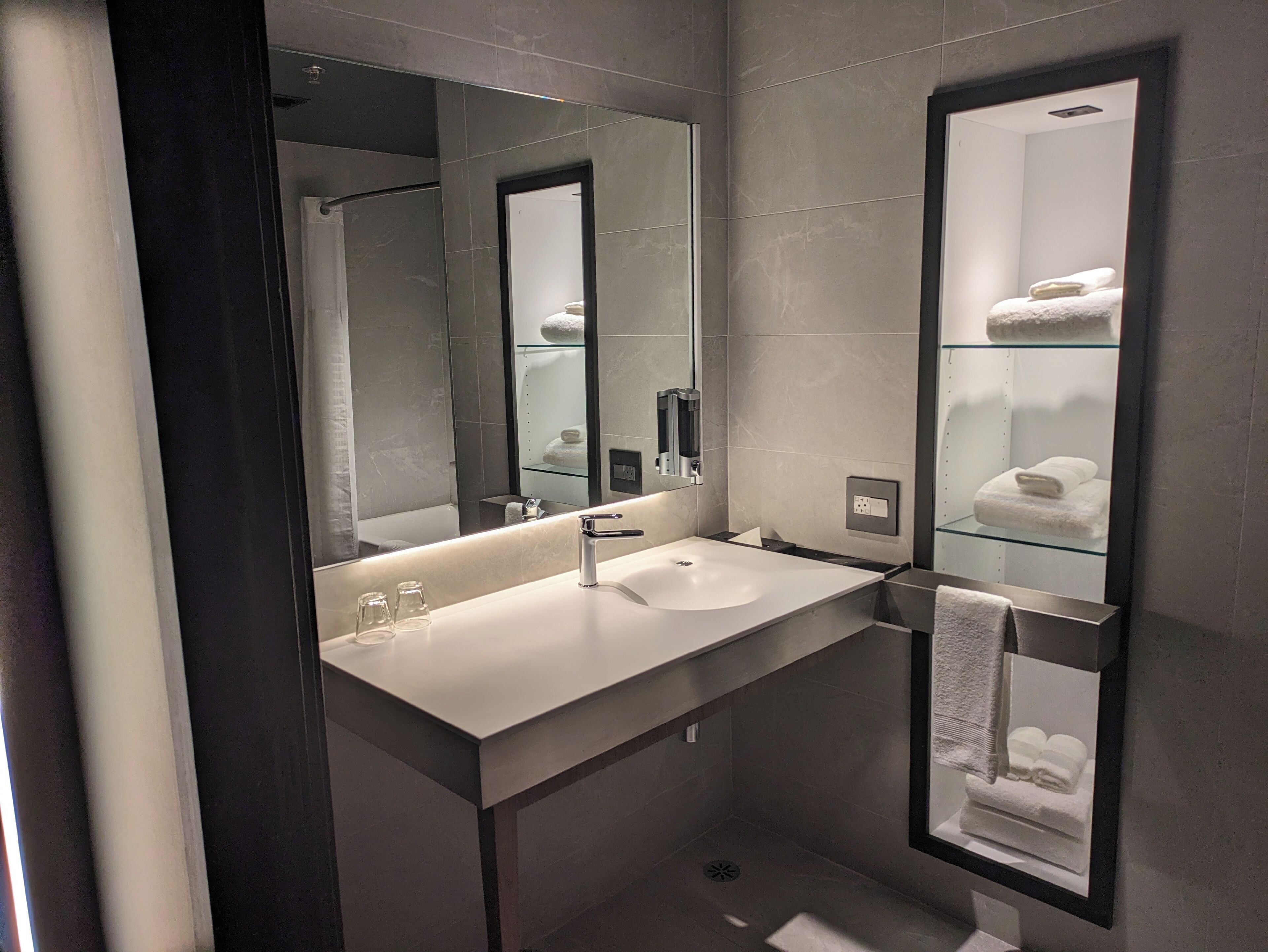 Un baño moderno con un espejo grande, un lavabo blanco y un estante de vidrio con toallas.