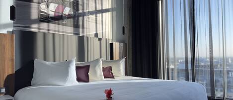 Una habitación de hotel moderna con una cama grande, una mesita de noche, un televisor empotrado en la pared que muestra un coche de carreras y vistas a la ciudad a través de amplias ventanas.