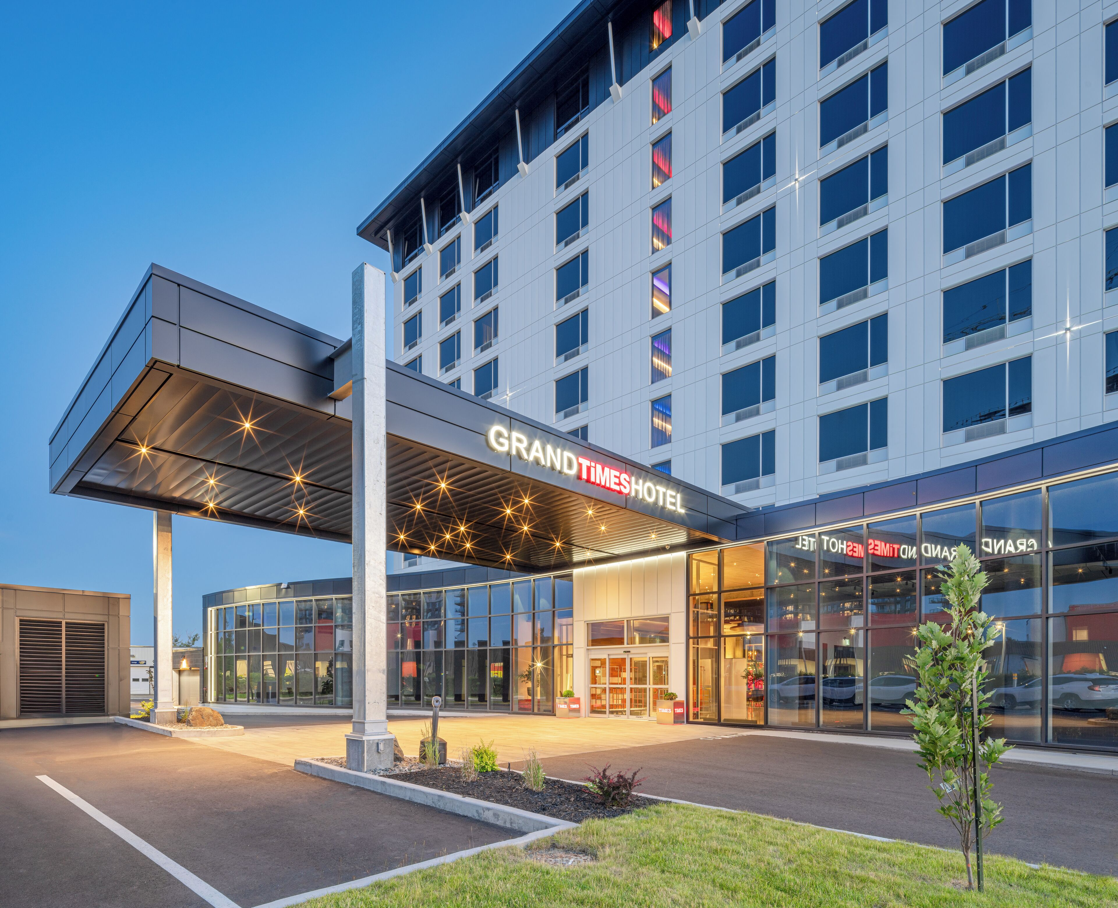 A modern hotel with a glass facade, a covered entrance, and a sign reading "GRANDTIMESHOTEL".