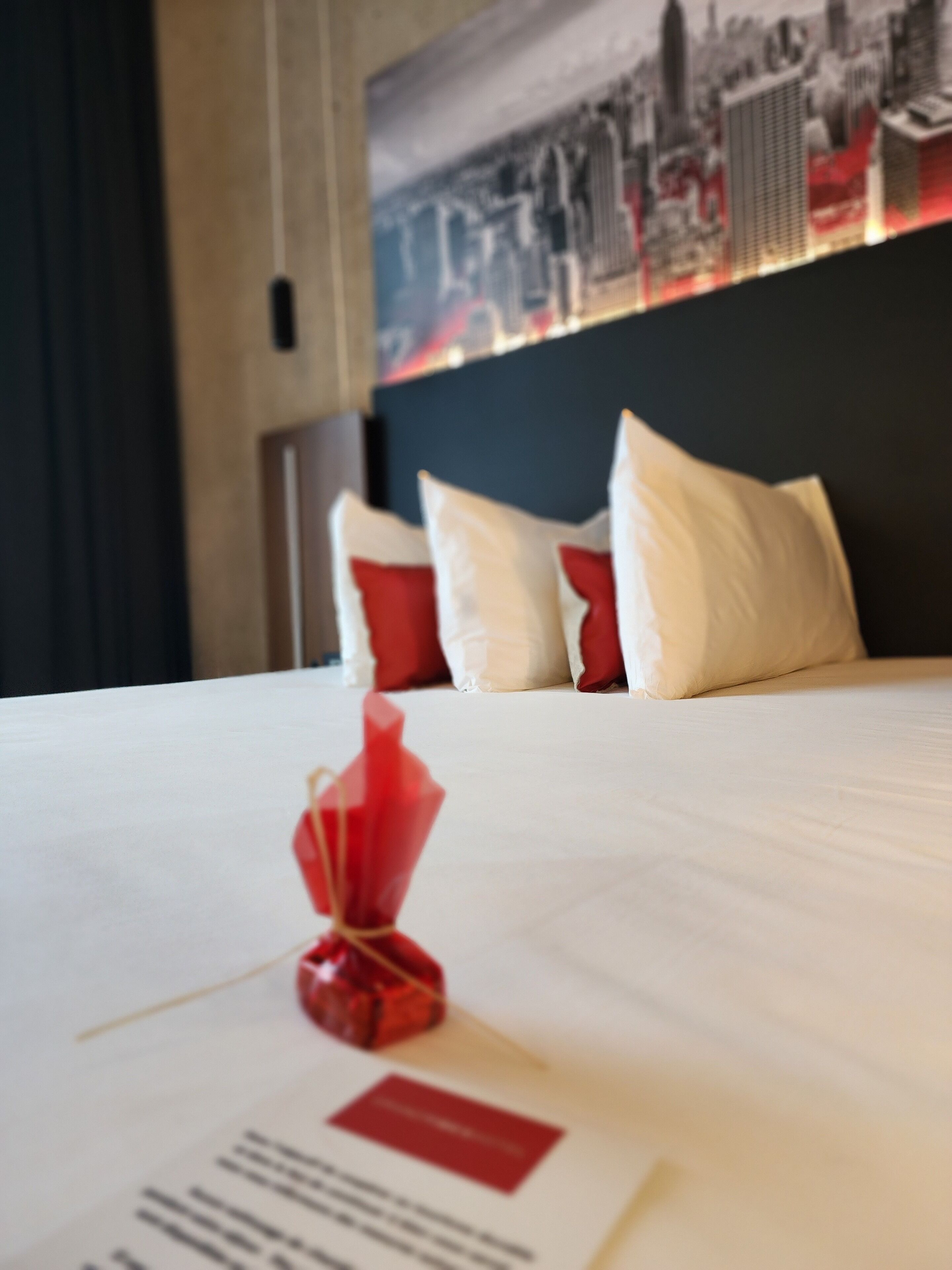 Une chambre d’hôtel avec un lit soigneusement fait, un cadeau rouge posé sur le lit et, au mur, une vue sur le paysage urbain.