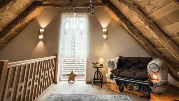 A cozy attic bedroom with a sloped ceiling, wooden beams, a large window, a black sofa, a small table with a lamp, and a patterned rug. - Ferienwohnung Josefine (Münster Albachten)