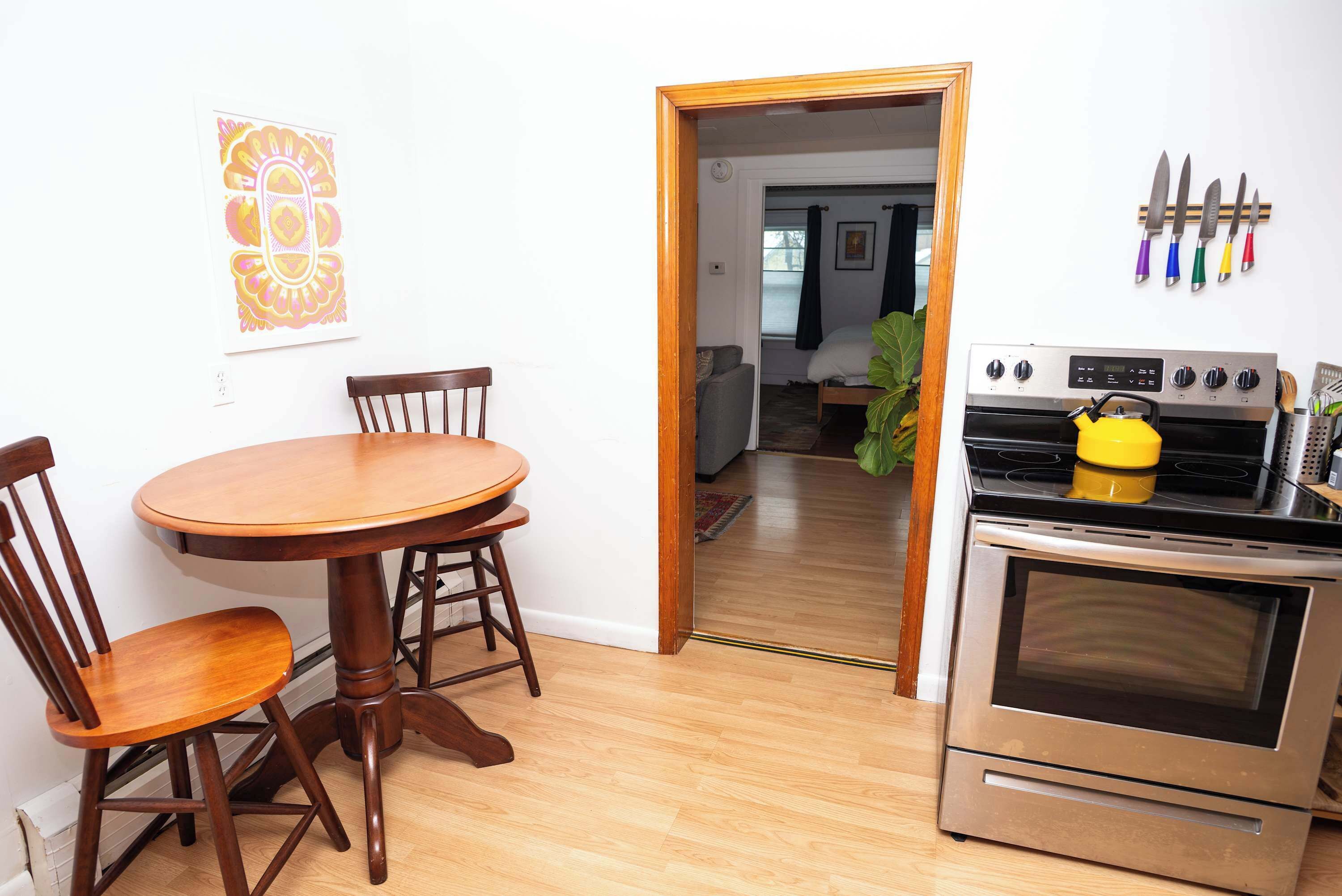 A dining area with a wooden table and chairs, a kitchen with an oven and a kettle, and a hallway leading to a bedroom.