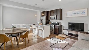 A modern kitchen with a central island, bar stools, a dining table with chairs, a TV, and a sofa in a living area.