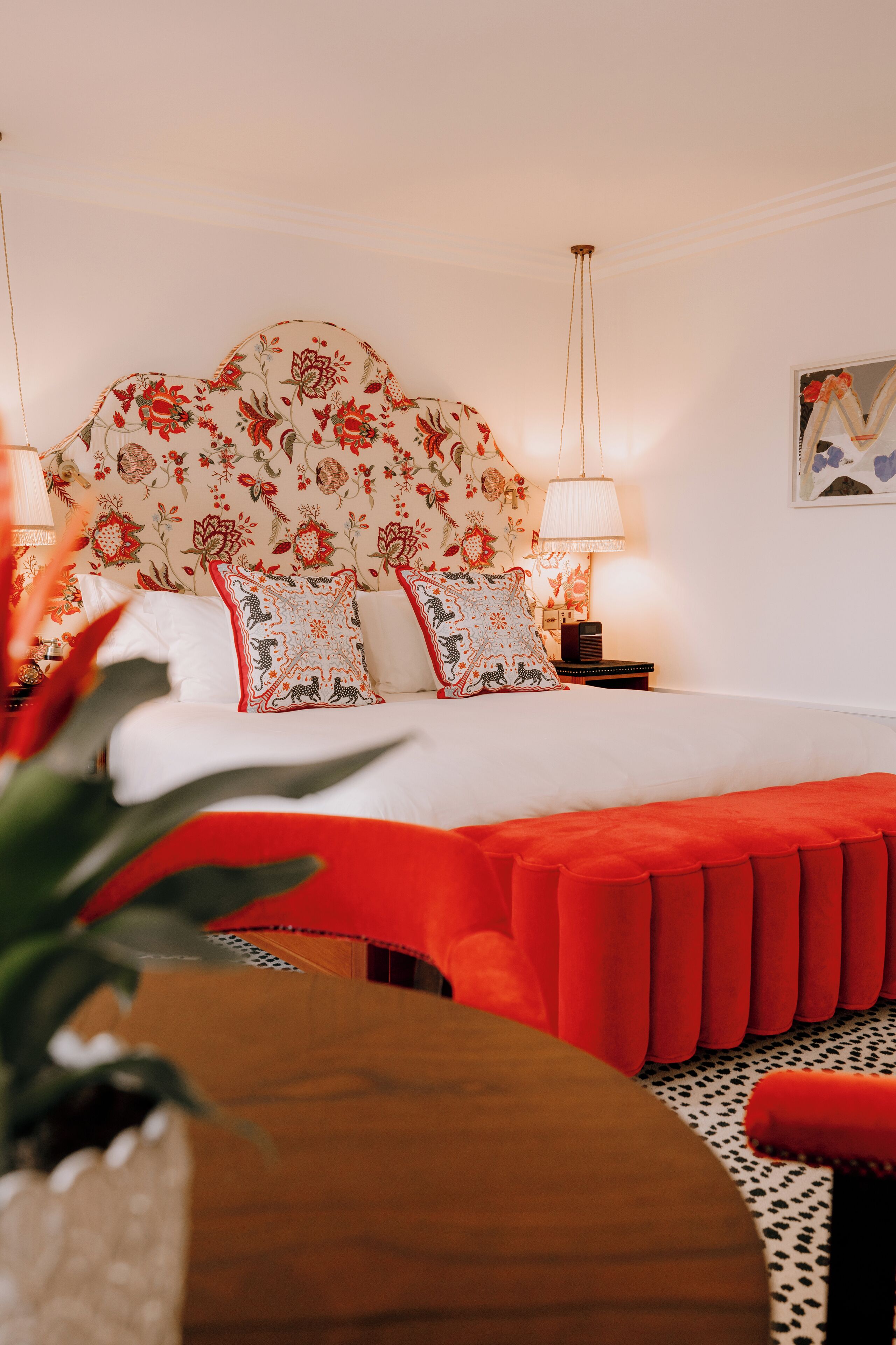 A hotel room with a floral-patterned headboard, a red accent bench, and a nightstand with a lamp.