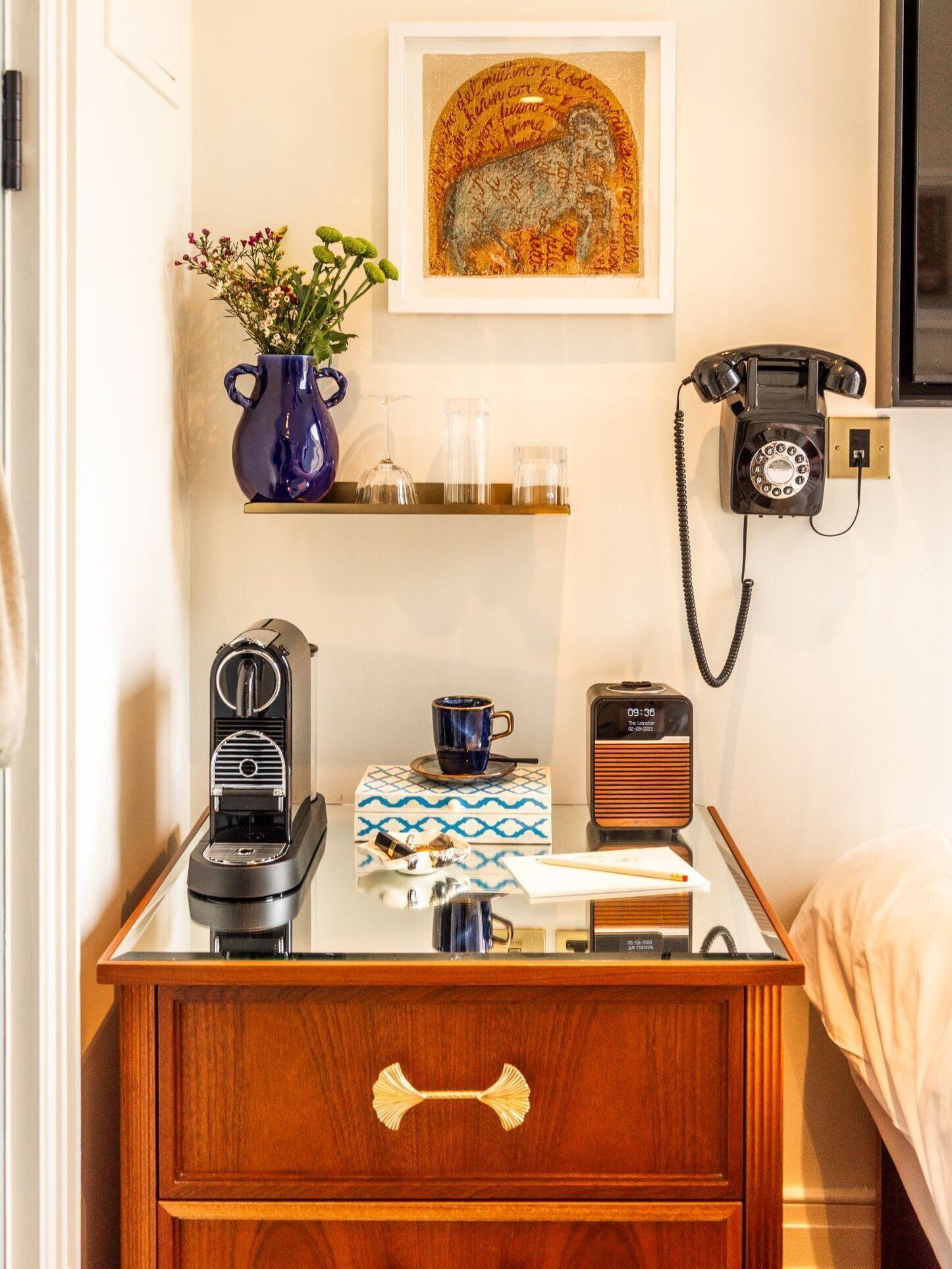 A neatly organized hotel room with a wooden nightstand, a vintage rotary phone, a radio, a coffee machine, a vase with flowers, and a framed artwork on the wall.