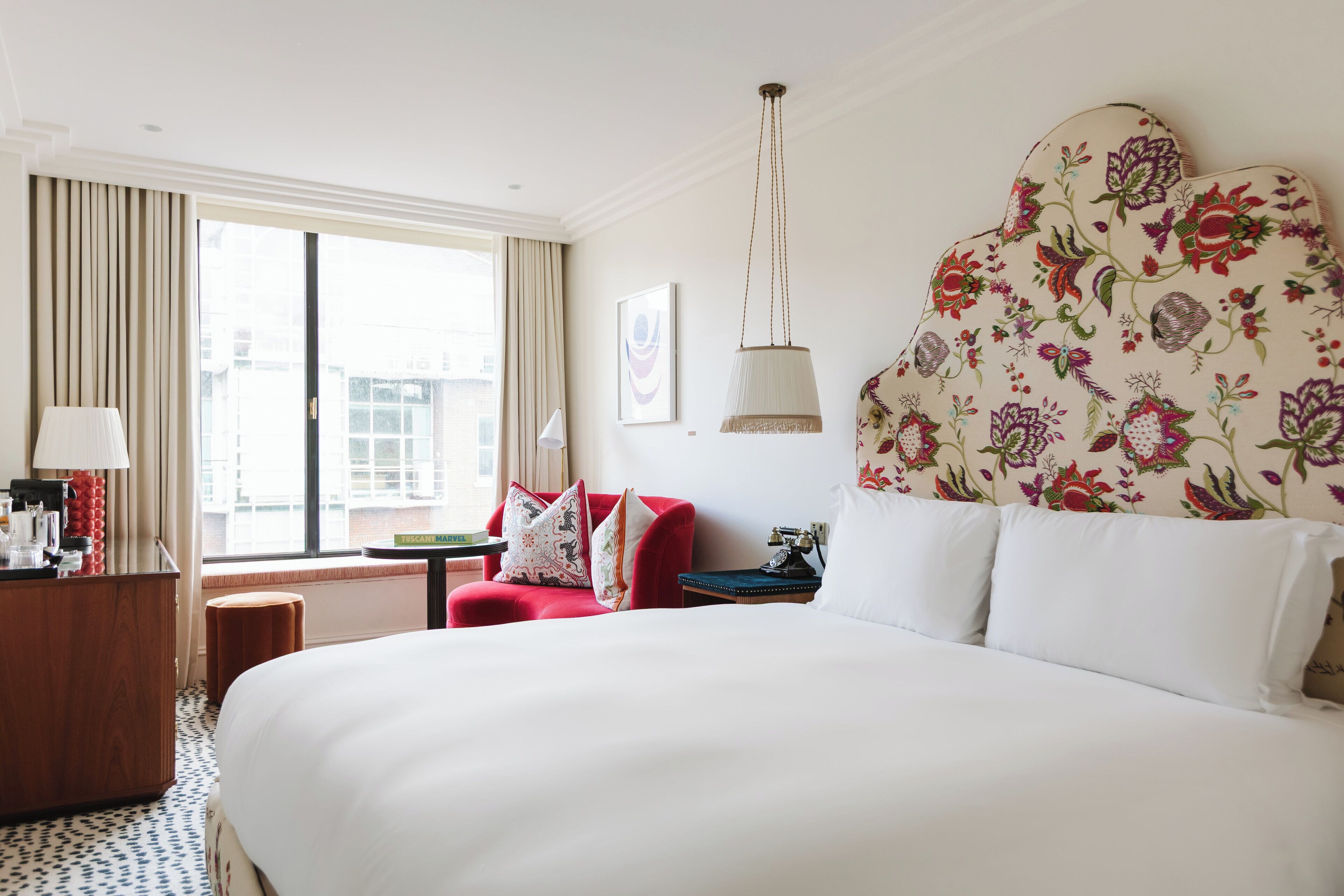 A hotel room with a large bed, a red armchair, a desk with a lamp, and a window with curtains.