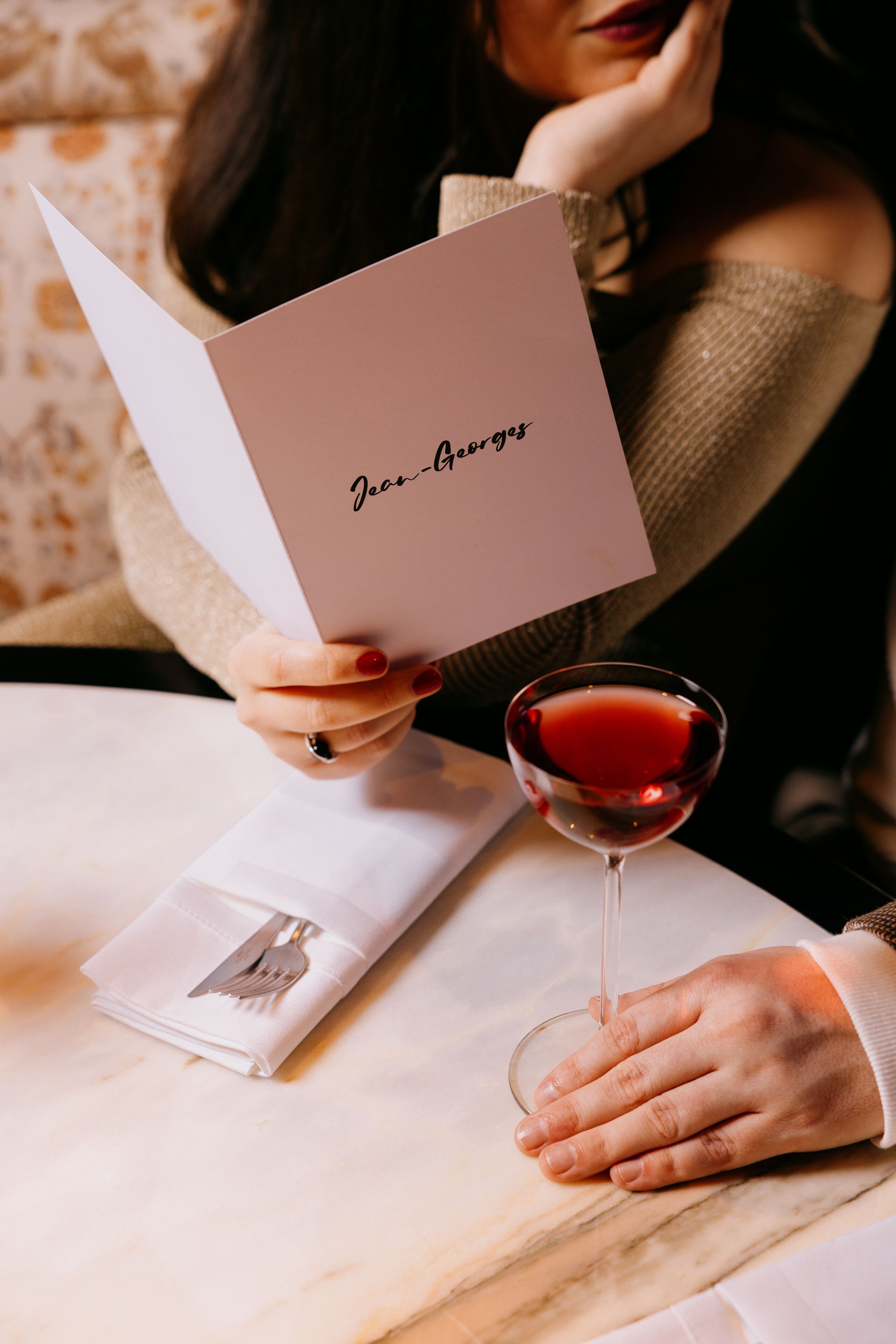 A person holding a card with "Jean-Guillaume" written on it, a folded napkin with a fork, and a glass of red wine on a table.