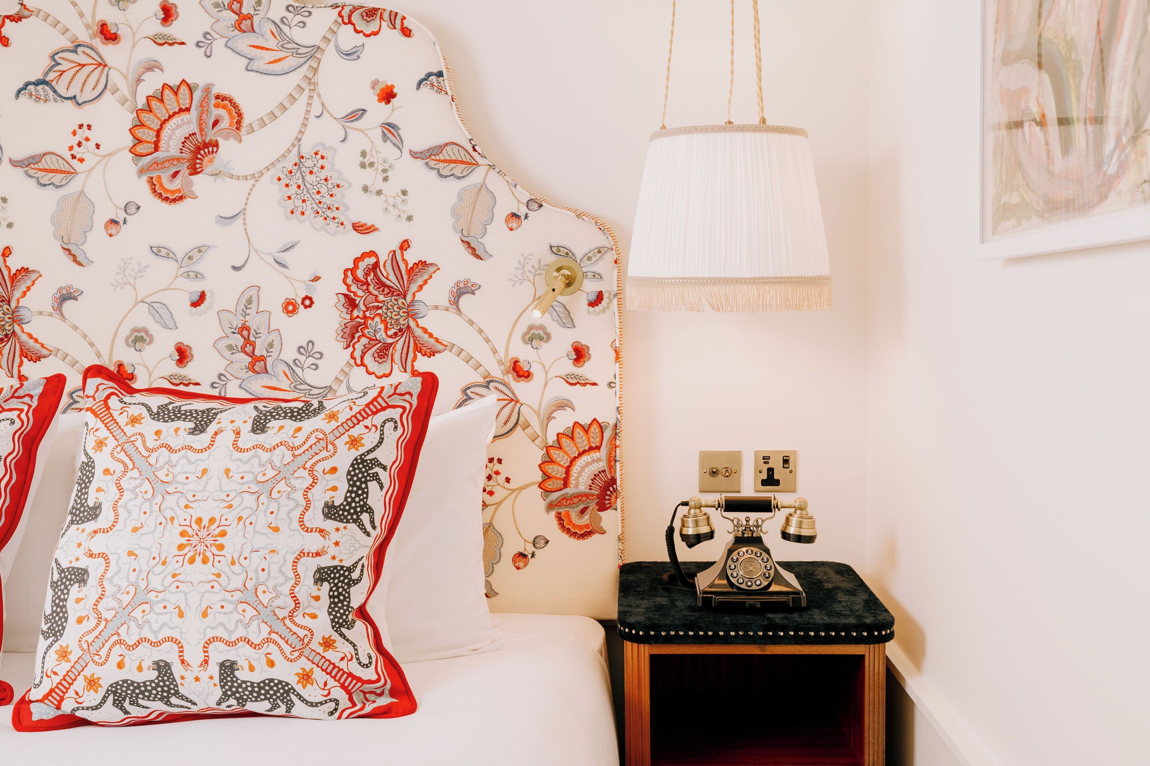 A room with a floral-patterned headboard, a white pillow with a red and black design, a vintage telephone on a wooden side table, and a hanging lamp.