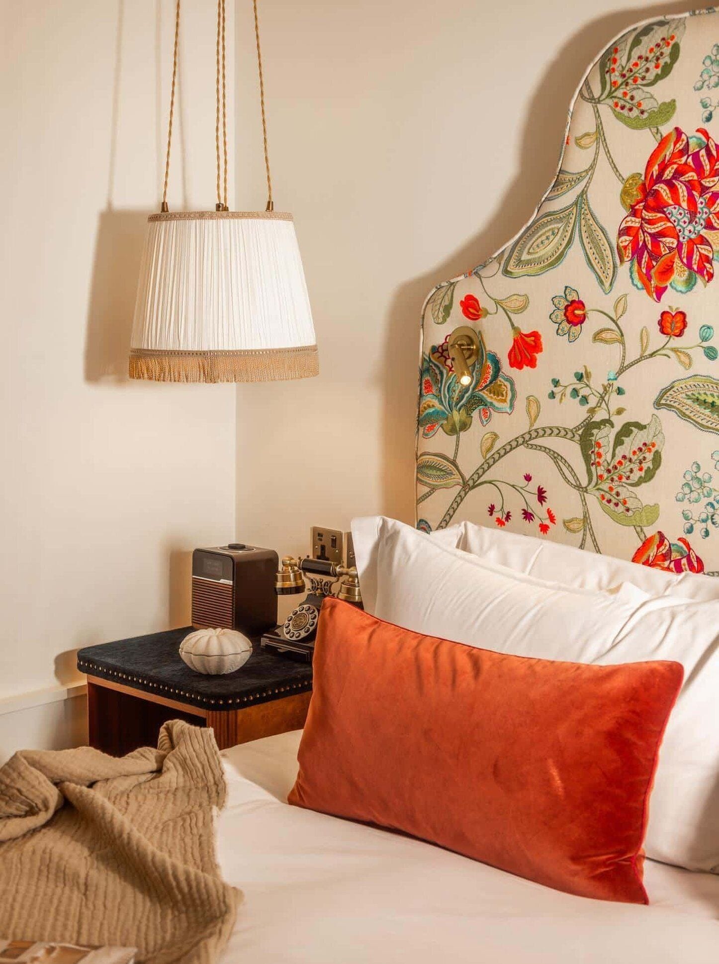 Een slaapkamer met een bed, nachtkastje, lamp en een hoofdeinde met bloemenprint.