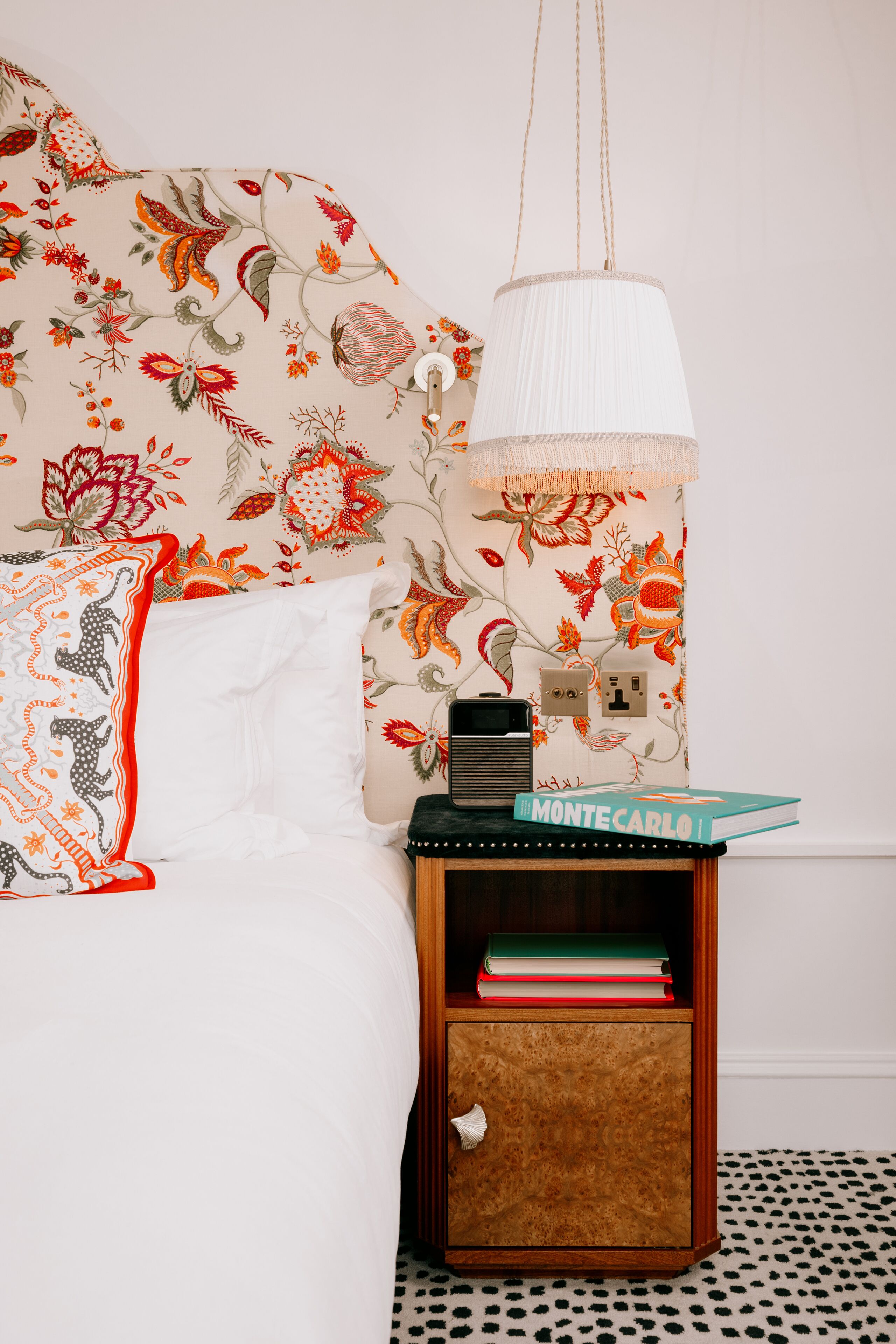 A bedroom with a floral wallpaper, a white bed, a bedside table with books, and a hanging lamp.