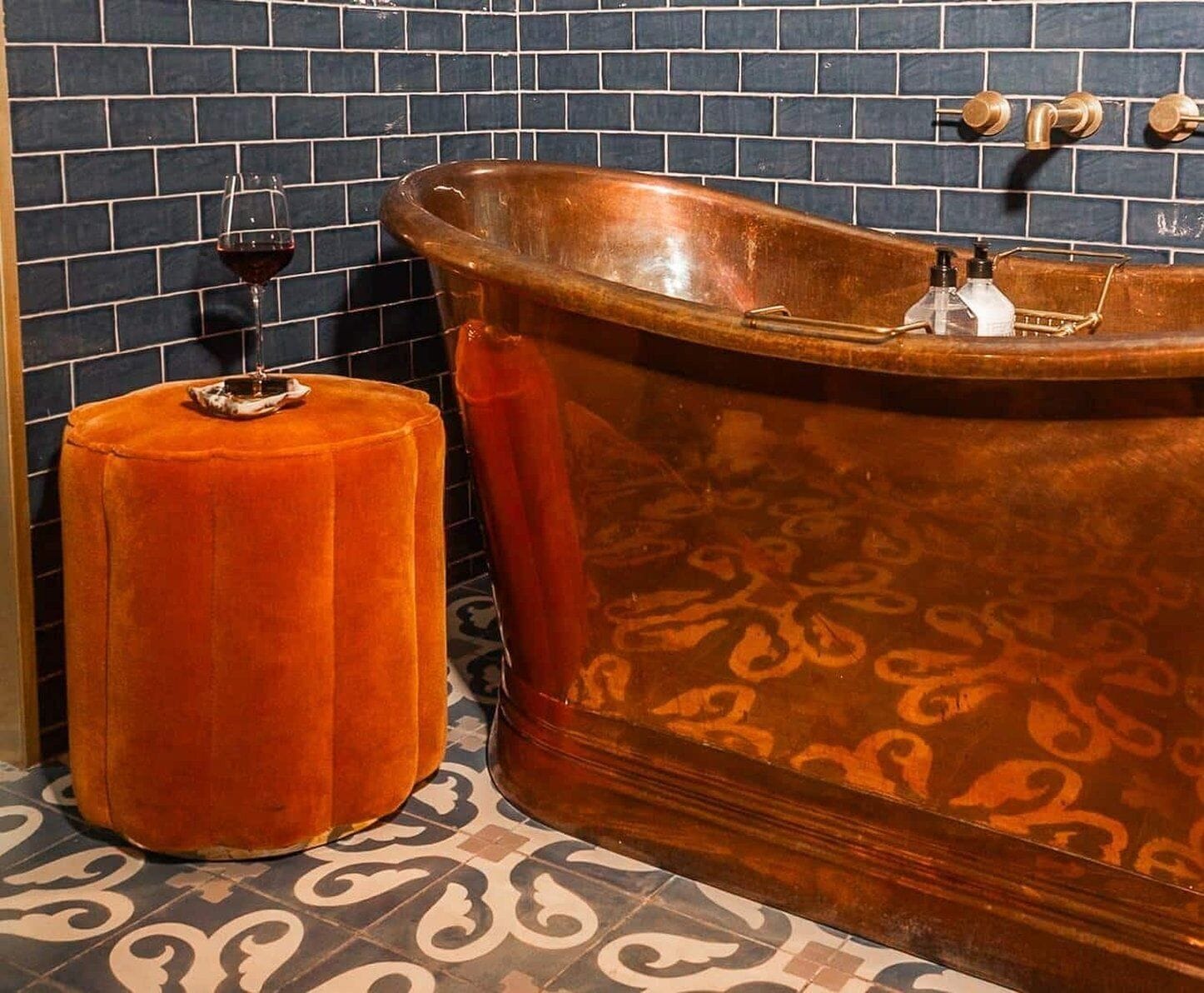 A bathroom with a copper bathtub, a round orange stool, and a wine glass on the floor.