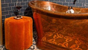 A bathroom with a copper bathtub, a round orange stool, and a wine glass on the floor.