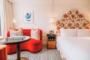 A hotel room with a red armchair, a small round table with a book, a bed with a floral headboard, and a nightstand with a lamp. - The Leinster (Dublin)
