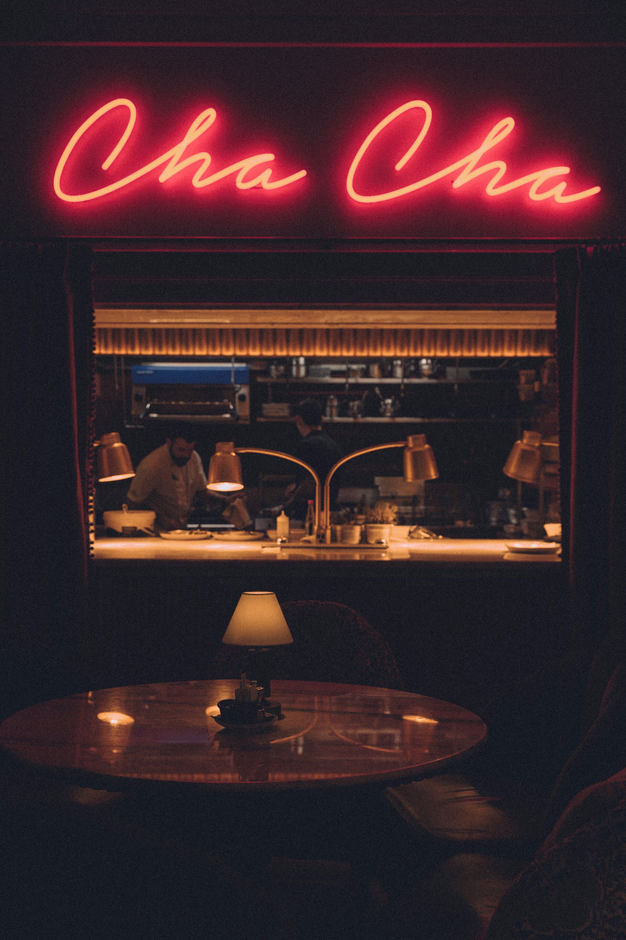 Ein schummrig beleuchtetes Restaurant mit einem Neonzeichen mit dem Schriftzug "Cha Cha" und Blick in die Küche durch ein Fenster.