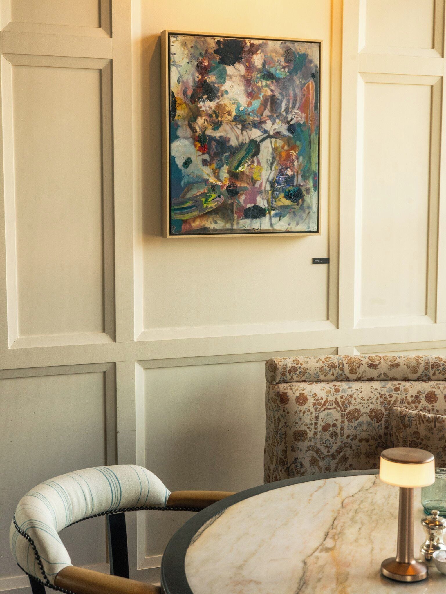 Una habitación con una mesa redonda de mármol, una silla con tapicería a rayas, un sofá estampado con motivos florales y un cuadro abstracto y colorido en la pared.