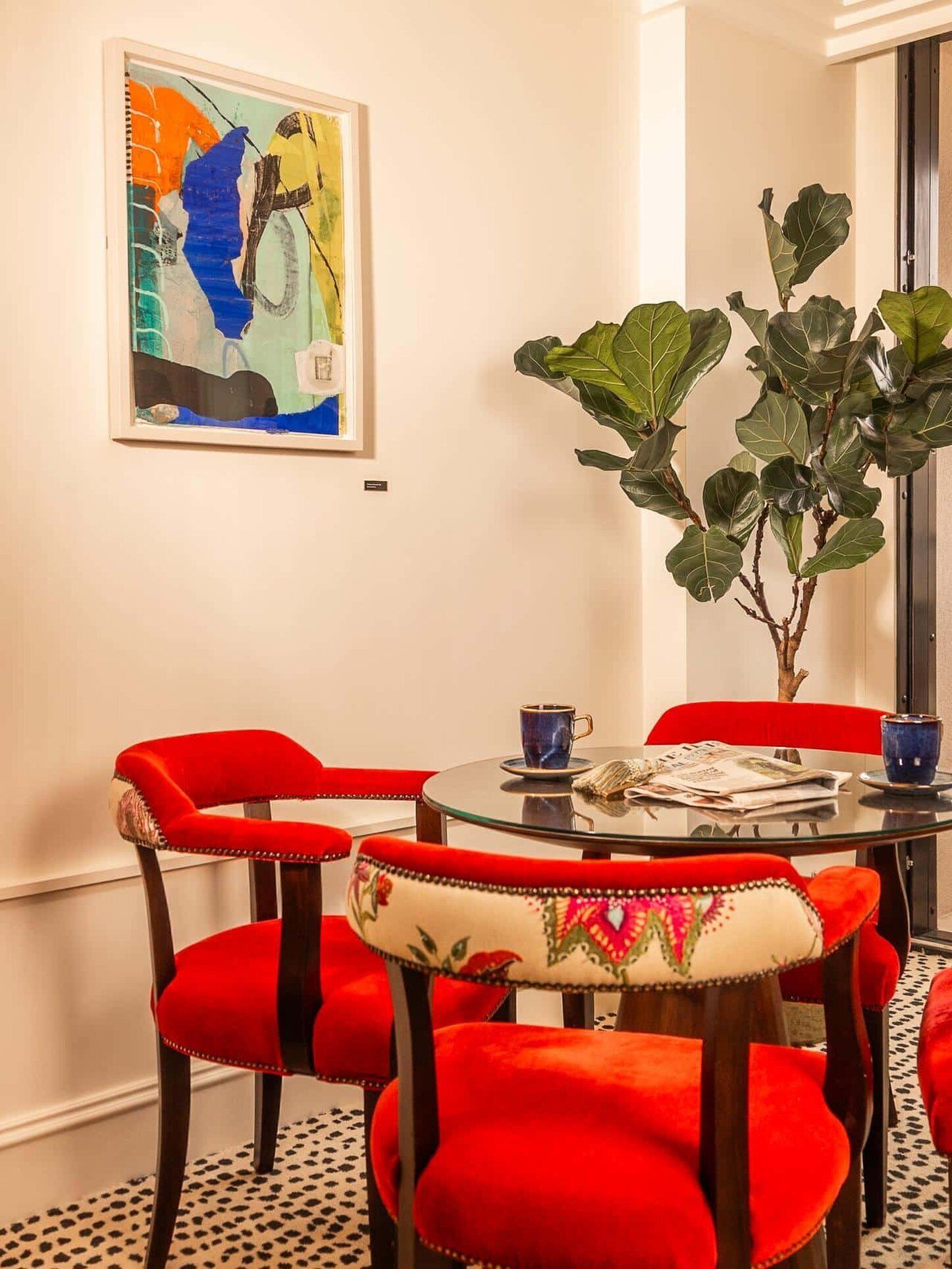 Un comedor con una mesa redonda, sillas tapizadas en rojo, una planta grande en maceta y un cuadro abstracto colorido en la pared.