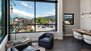A modern hotel room with a large window offering a view of a street and buildings. There are two leather sofas, a dining table with chairs, and a small side table with bottles.