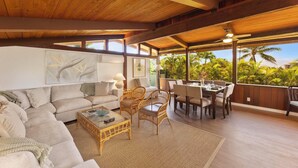 A spacious living area with a wooden ceiling, a sofa, a coffee table, wicker chairs, a dining table with chairs, and large windows overlooking a tropical landscape.