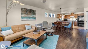 Ein modernes Wohnzimmer mit einer Couch, einem Couchtisch und Sesseln, eine Essecke mit Tisch und Stühlen sowie eine Küche mit Holzschränken und einem Kühlschrank.