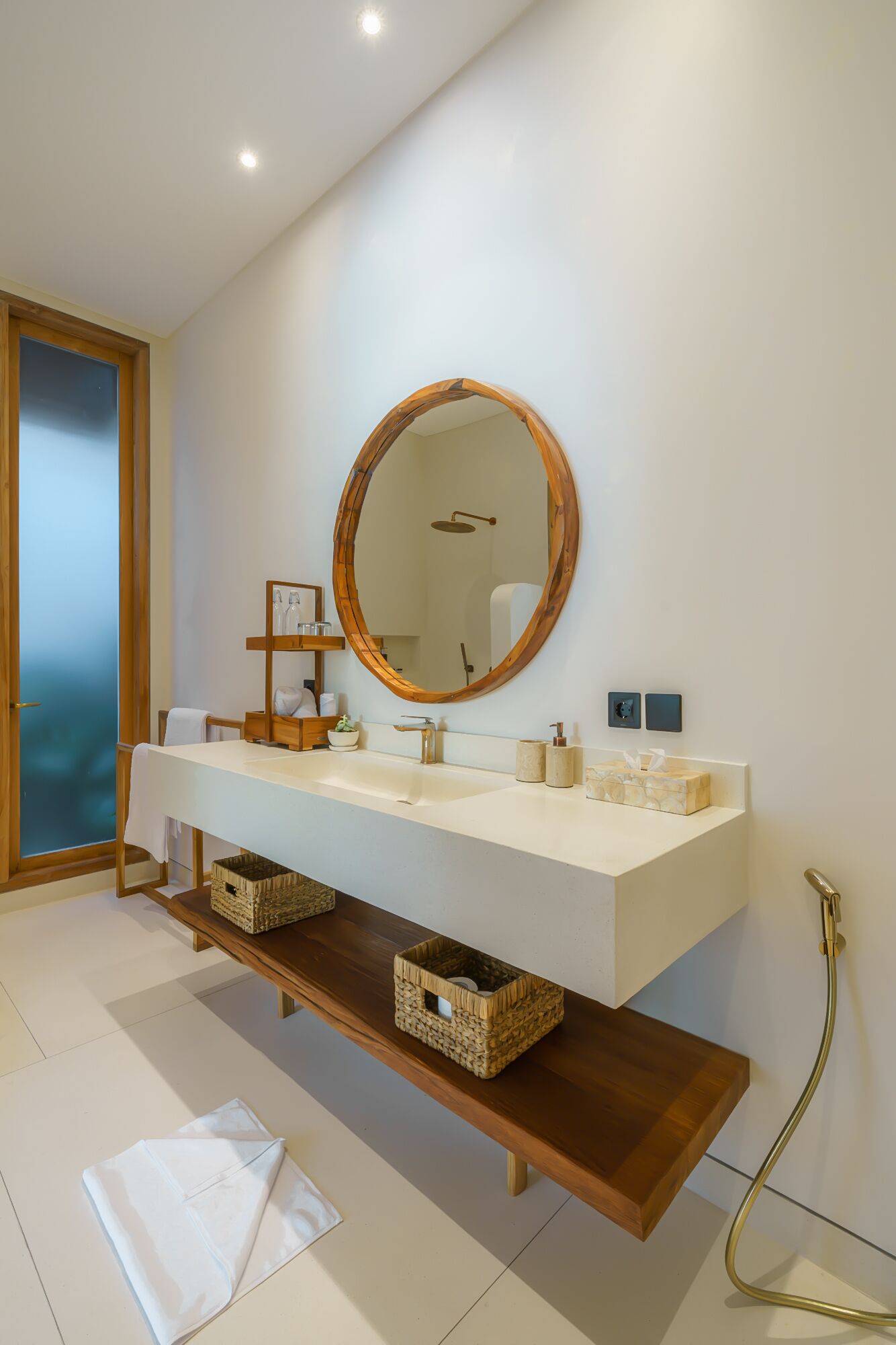 A modern bathroom with a white vanity, a round mirror, and a wooden shelf.