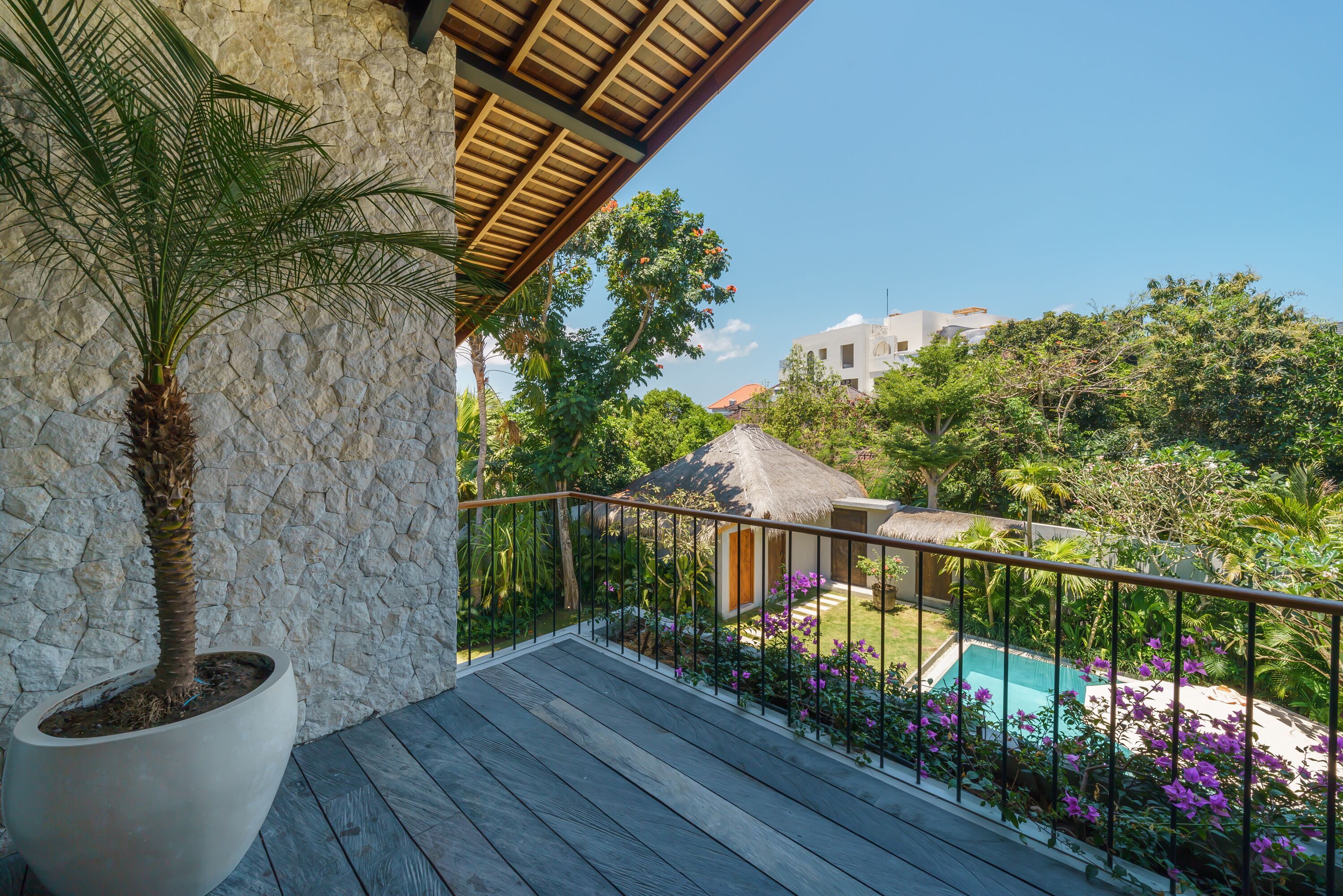 A wooden deck with a stone wall, a potted palm tree, and a view of a swimming pool surrounded by purple flowers.