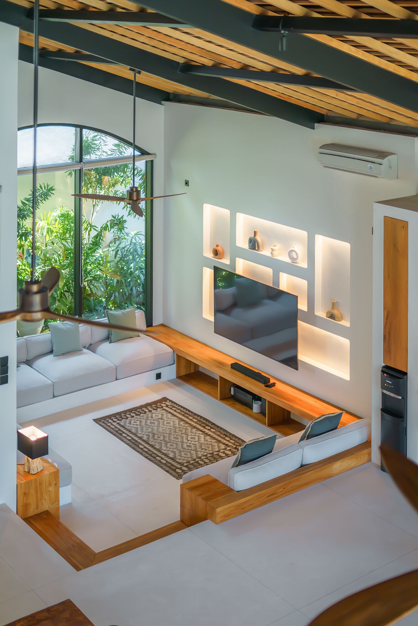 A modern living room with a large flat-screen TV, built-in shelves with decorative items, a wooden bench, and a view of greenery through large windows.