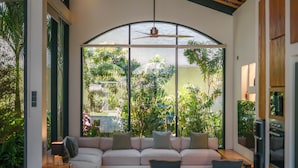 A modern living room with a large sectional sofa, a ceiling fan, and a view of lush greenery outside. - Casa Donilo  (Canggu)
