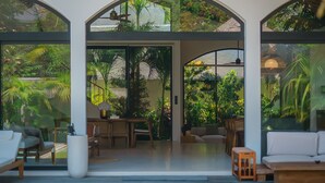 A modern, open-plan living area with a dining set and seating, large windows, and a view of lush greenery. - Casa Donilo  (Canggu)
