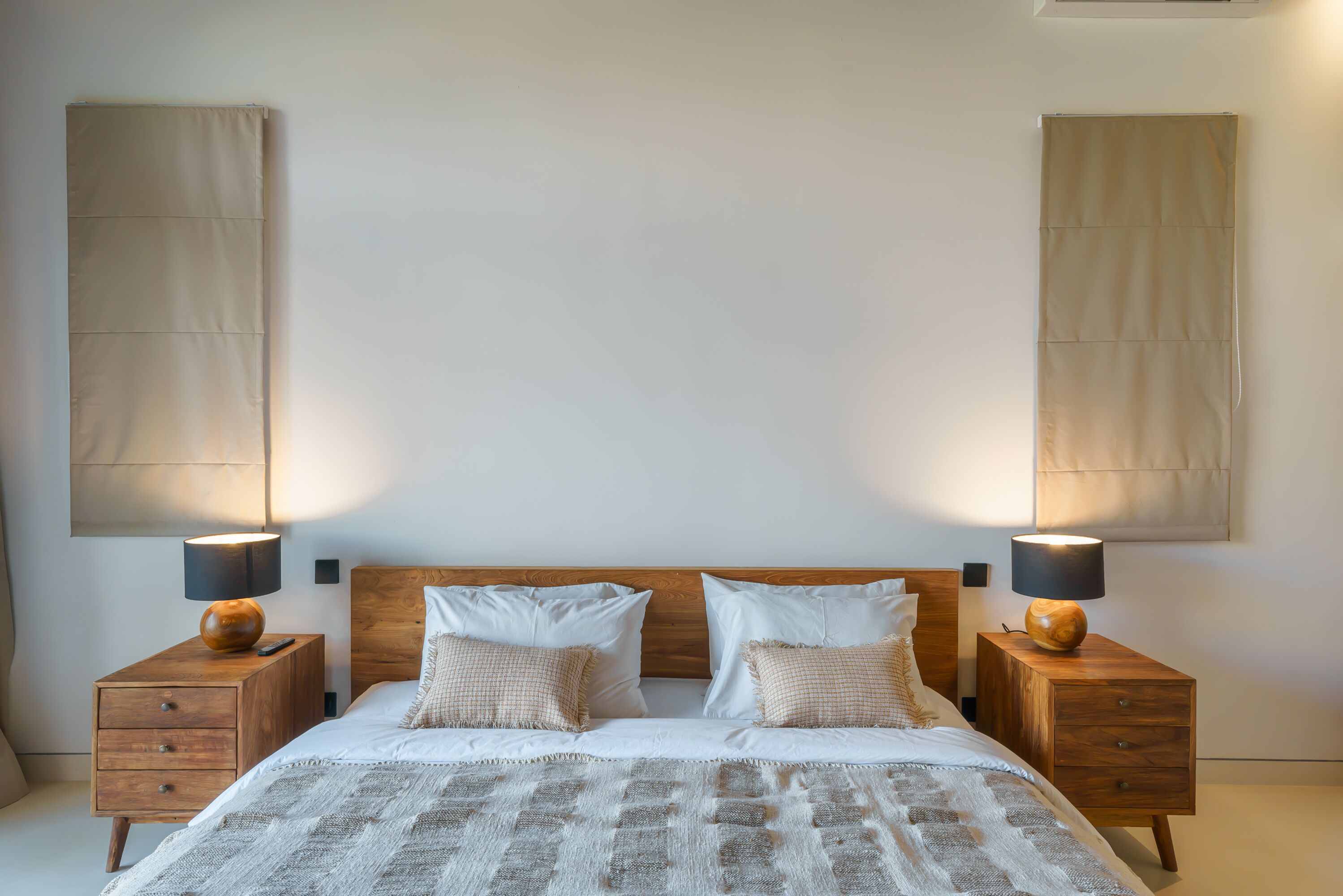 Un dormitorio con cabecera de madera, dos mesitas de noche con lámparas, cama con ropa de cama blanca y una manta decorativa.