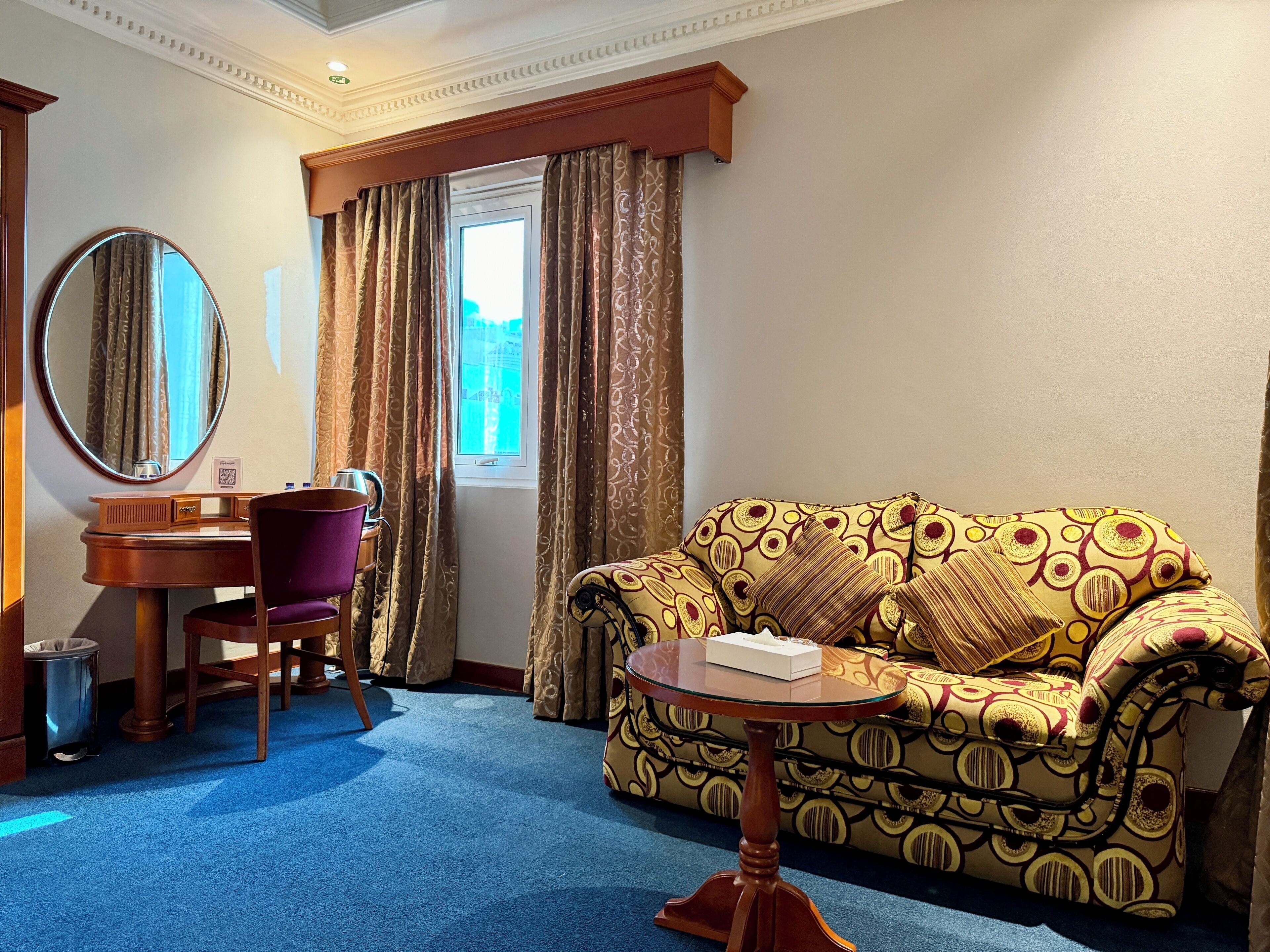 Ett hotellrum med ett blått golv, en rund spegel, ett skrivbord i trä med en lila stol, en mönstrad soffa och ett fönster med gardiner.