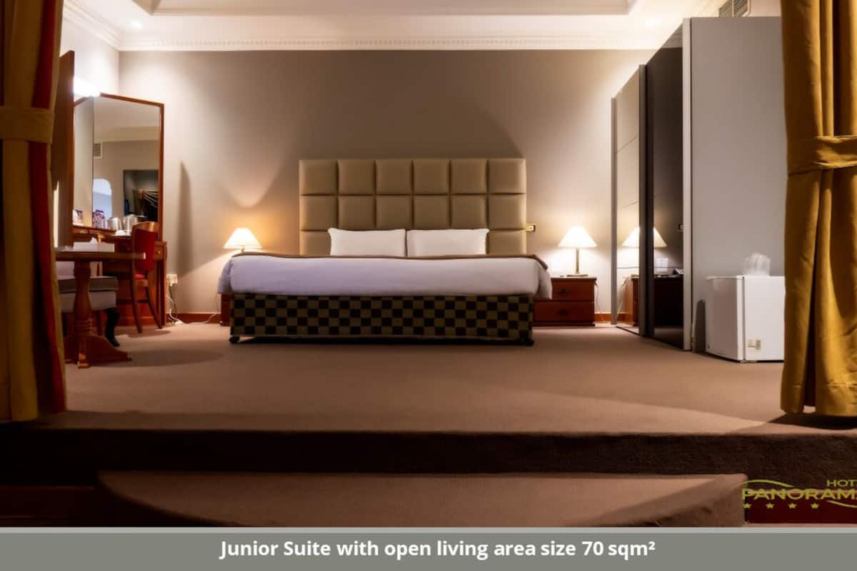 A junior suite with an open living area size of 70 sqm², featuring a large bed, bedside tables, a desk, and a chair.