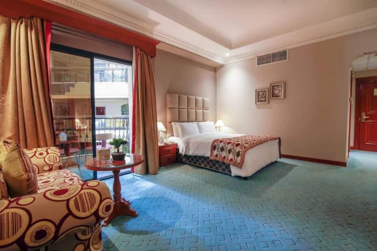 A hotel room with a bed, a sofa, a small table with a vase, and a window with curtains.