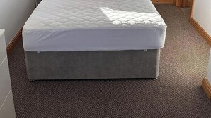 Una cama bien hecha con un colchón acolchado en una habitación con ventana, cortinas, una mesita de noche con un ventilador y una puerta de madera.