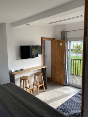 A modern bedroom with a bed, a desk with two stools, a flat-screen TV, and a sliding glass door leading to a balcony. - F17 Rickardos Holiday Lets (Mablethorpe)