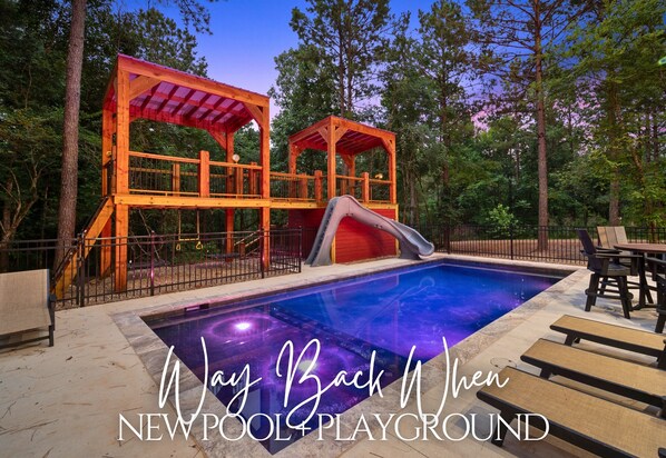 A wooden play structure with a slide and a red-roofed pavilion, surrounded by a swimming pool and trees.