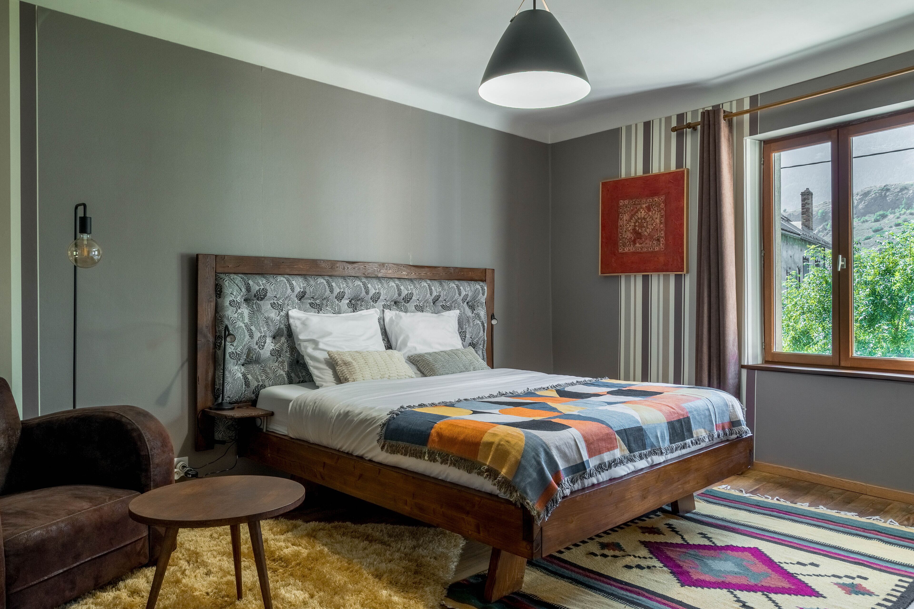 A bedroom with a wooden bed, a small round table, a fluffy rug, a patterned rug, a hanging light fixture, and a window with curtains.