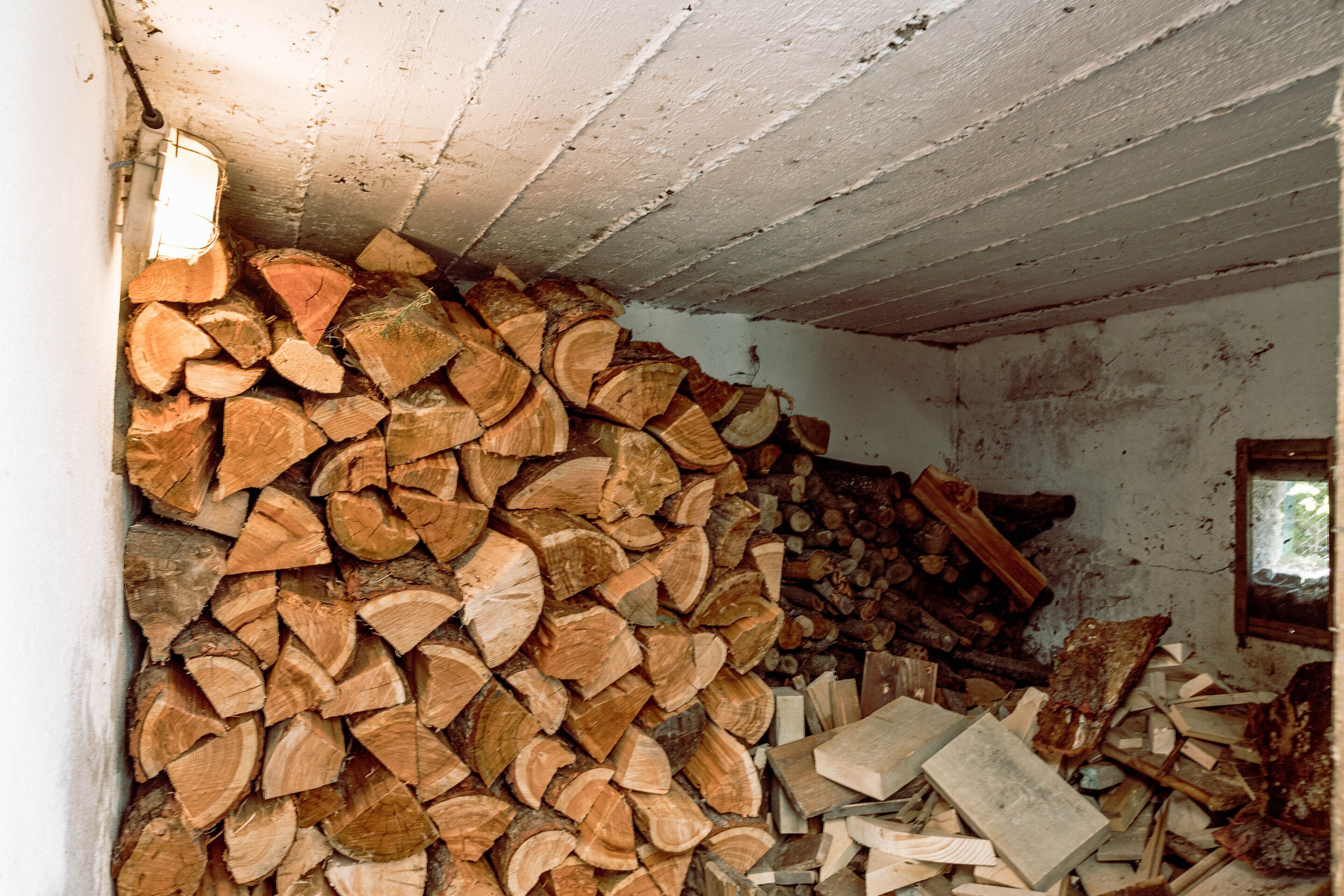 A room with a large stack of firewood against the wall and a window with a view of greenery outside.