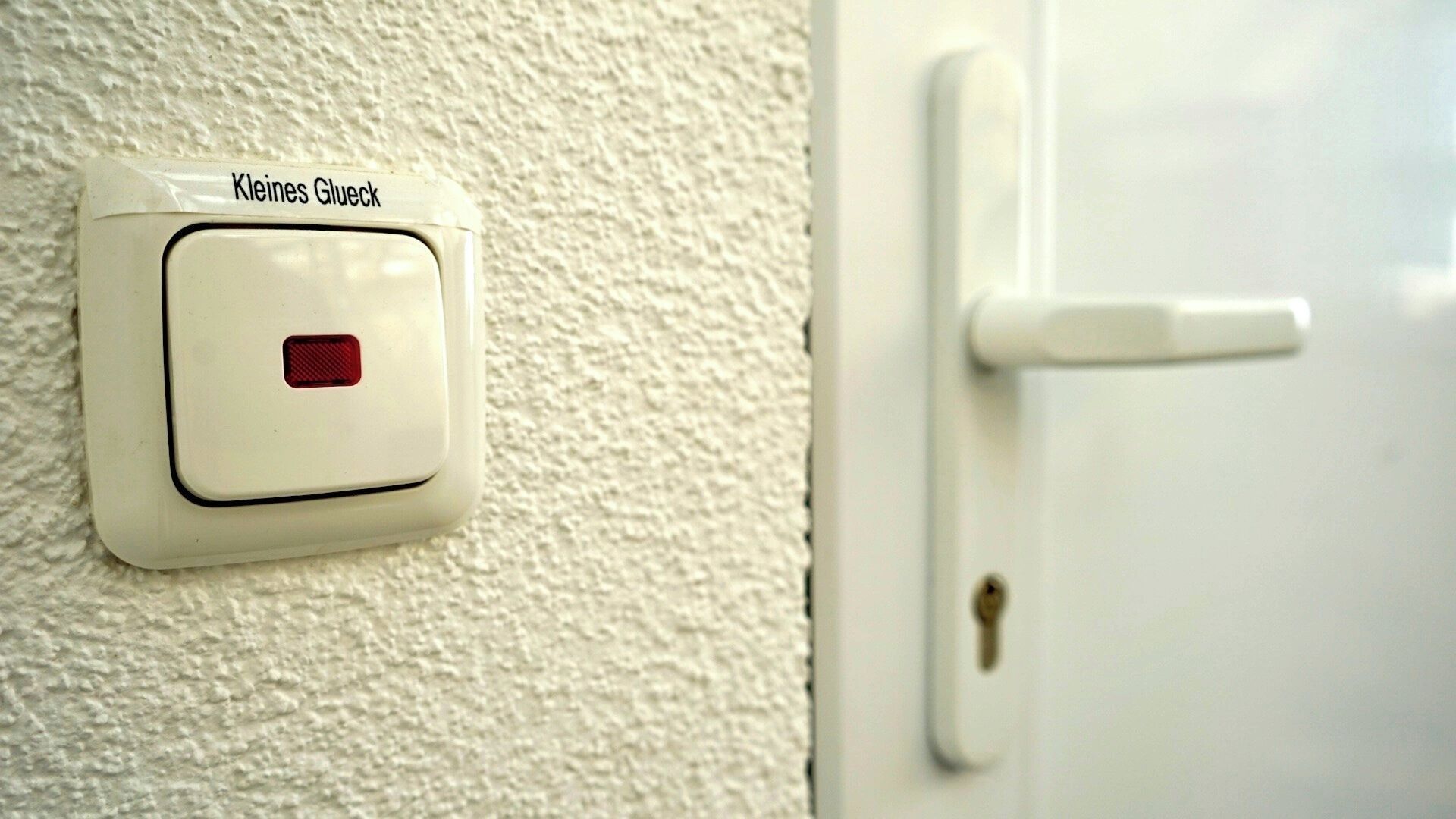 A white door with a keyhole and a wall-mounted alarm button labeled "Kleines Glueck".